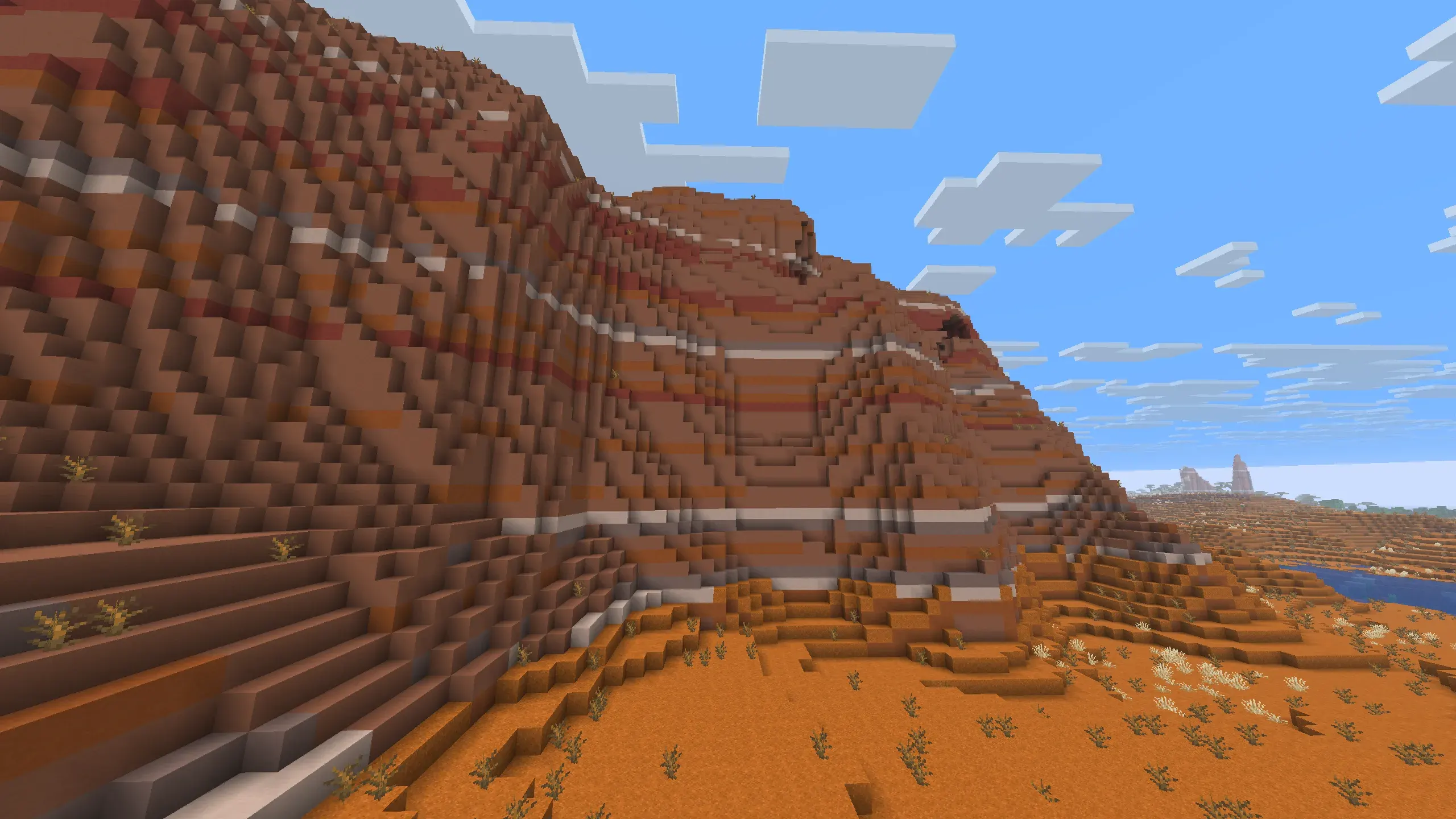 Minecraft Badlands biome with massive layered terracotta mountain, orange sand valley, scattered dead bushes, and distant mesas under a bright blue sky
