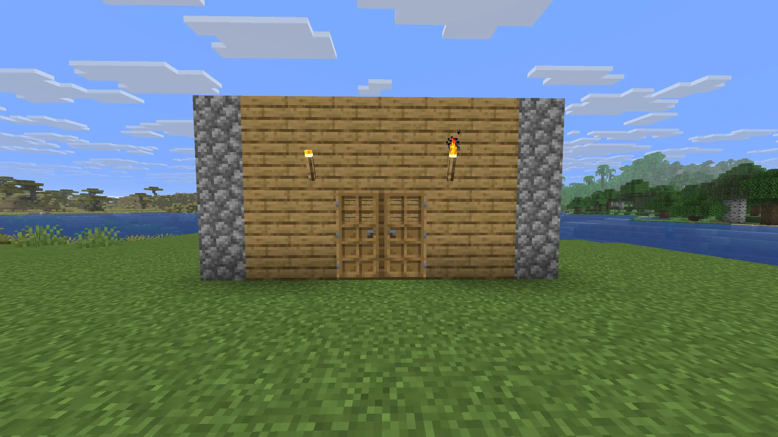 Front view of a small Minecraft starter base made of oak wood planks with cobblestone pillars on the sides, featuring double wooden doors and lit torches mounted on the wall, situated next to a river.